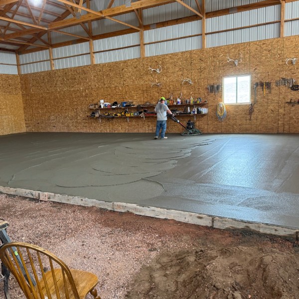 Worker smoothing a freshly poured concrete floor inside a wooden-frame agricultural building.