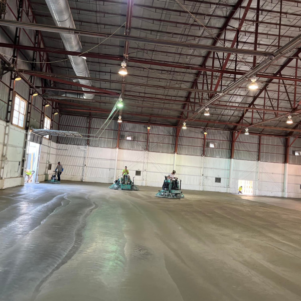 Workers smoothing a freshly poured concrete floor inside a large industrial warehouse.