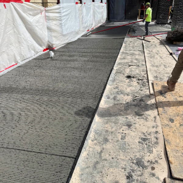 Worker creating a textured finish on a freshly poured concrete sidewalk beside a commercial building.